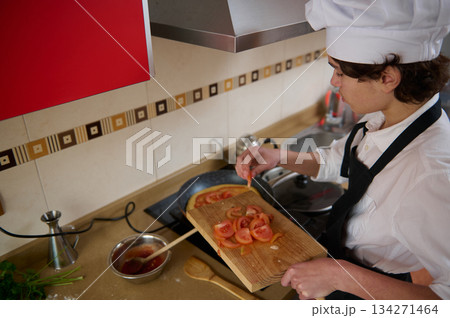 Culinary Student In White Hat And Apron Slices Tomatoes On Wooden Board In Modern Kitchen Culinary Student In White Hat And Apron Slices Tomatoes On Wooden Board In Modern Kitchen 134271464