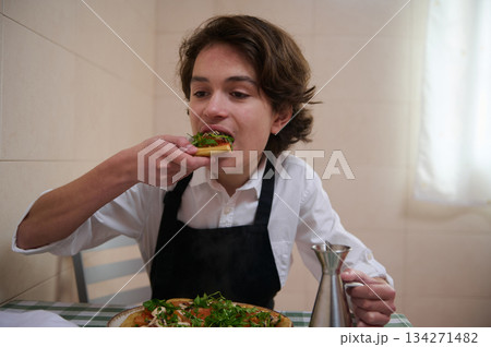 Young Man in White Shirt and Apron Eating Fresh Pizza in Kitchen Young Man in White Shirt and Apron Eating Fresh Pizza in Kitchen 134271482
