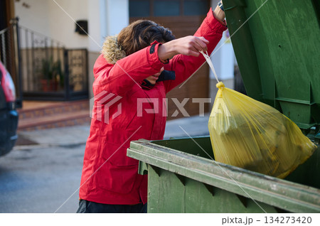 Person In Red Jacket Disposes Yellow Trash Bag Into Green Dumpster On Street 134273420