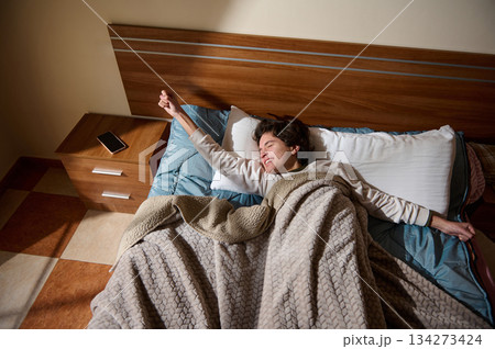 Young Man Laughing And Stretching In Bed With Cozy Blanket In Bedroom Morning Young Man Laughing And Stretching In Bed With Cozy Blanket In Bedroom Morning 134273424