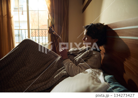 Young Boy Relaxing in Bed with Blanket, Reading on Tablet by Window Light 134273427