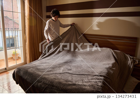 Person Makes Bed In Cozy Bedroom With Blanket, Sunlight, And Striped Wall Decor 134273431