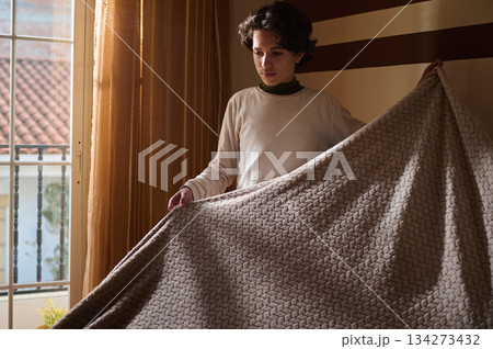 Young Man Unfolds Cozy Blanket In Sunlit Room For Comfort And Warmth Young Man Unfolds Cozy Blanket In Sunlit Room For Comfort And Warmth 134273432