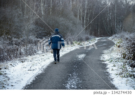 Senior caucasian man walking alone along a snowy rural road in winter forest. Concept of solitude, journey, healthy lifestyle and active aging. 134274430