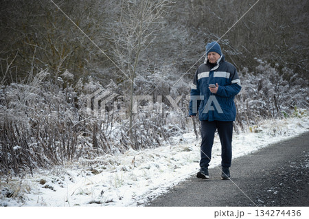Winter walk. Adult man checking smartphone while walking outdoors in publick park. Concept of modern lifestyle, technology, communication and active senior age. 134274436