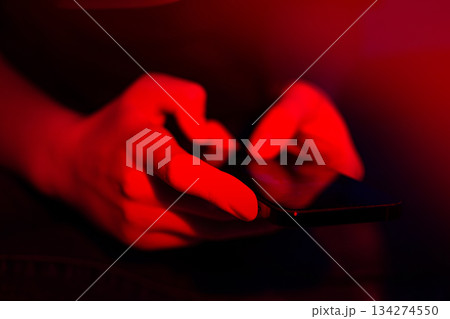 Cropped woman hand using smartphone on a neon glowing background. Happy young girl looking at screen close. Cropped woman hand using smartphone on a neon glowing background. Happy young girl looking at screen close. 134274550