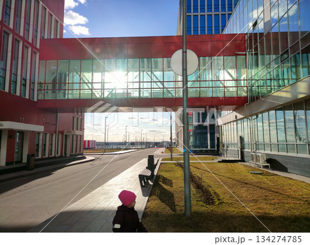 Child walking on a sunny day past modern office buildings with a connecting red glass skywalk, featuring contemporary architecture and urban infrastructure under a clear blue sky. Child walking on a sunny day past modern office buildings with a connecting red glass skywalk, featuring contemporary architecture and urban infrastructure under a clear blue sky. 134274785