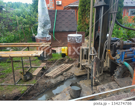 A drilling rig works in a mud trench, creating a well for a domestic water supply. Pipes and construction equipment for drilling the well are visible at the construction site. 134274786