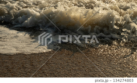 An ocean wave crashes onto wet sand, white sea foam catching golden rays of sunlight and reflecting warm hues. A close-up coastal seascape evokes a sense of calm and natural tranquility. 134274803