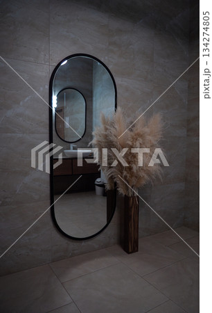 Modern bathroom interior with an oval mirror above a vanity and sink, neutral tiled walls and floor, and a tall vase of fluffy pampas grass for a soft, chic look. 134274805