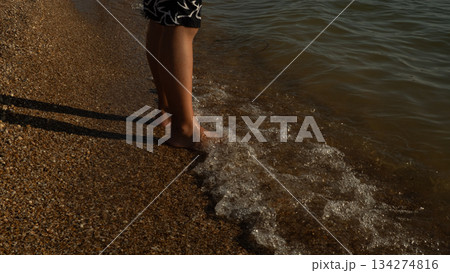 A person standing at the water's edge on a pebble beach feels the waves washing over their bare feet, creating a moment of oneness with nature in summer. Waves washing over a person's feet at sunset. 134274816