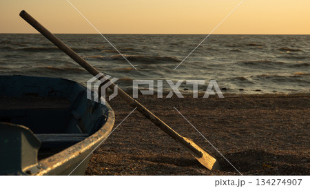 An old wooden oar balances on a small blue rowboat resting on the beach during the calm golden hour. Against the backdrop of calm waves, the old boat creates a serene and tranquil coastal landscape. 134274907