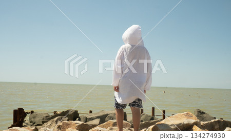 A teenager stands on a rocky seashore wearing a light hooded shirt and patterned swim shorts. He gazes out at the calm, sunlit sea under a clear summer sky, pensive and free. A teenager stands on a rocky seashore wearing a light hooded shirt and patterned swim shorts. He gazes out at the calm, sunlit sea under a clear summer sky, pensive and free. 134274930