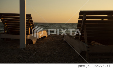 Sun loungers against the backdrop of a calm sea at sunset. Empty wooden sun loungers on a pebble beach invite relaxation and gazing at the calm sea under a warm, golden sunset sky. Sun loungers against the backdrop of a calm sea at sunset. Empty wooden sun loungers on a pebble beach invite relaxation and gazing at the calm sea under a warm, golden sunset sky. 134274931