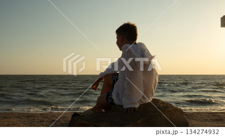 A boy sits on a coastal rock during golden hour, quietly and thoughtfully gazing at the ocean horizon. A teenager sits on the seashore at sunset, serene and peaceful, pensively gazing at the waves. A boy sits on a coastal rock during golden hour, quietly and thoughtfully gazing at the ocean horizon. A teenager sits on the seashore at sunset, serene and peaceful, pensively gazing at the waves. 134274932