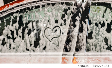 A car window covered in heart-shaped foam. A playful romantic gesture drawn in the foam during a car wash. Soft reflections of trees and sky, textured bubbles on the glass. 134274983