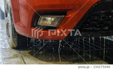 A wet car bumper with dripping water, shiny orange paint, and headlights. A car wash with pressure water creates dynamic splashes and soap streaks. A close-up of the car's bumper at the car wash. 134275040