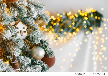 Festive Christmas tree lights ornaments small house and baubles on snowy branch on bokeh background Festive Christmas tree lights ornaments small house and baubles on snowy branch on bokeh background 134275181