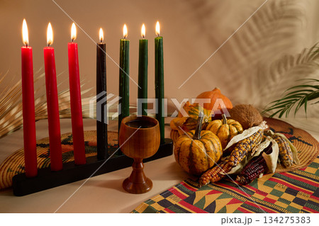 Kwanzaa Celebration Setting. Warm Kwanzaa scene with lit candles, harvest vegetables, and a wooden chalice Kwanzaa Celebration Setting. Warm Kwanzaa scene with lit candles, harvest vegetables, and a wooden chalice 134275383