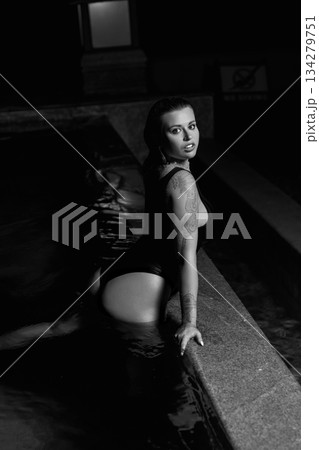 A sexy girl in a black swimsuit poses at night by the pool at a party. 134279751