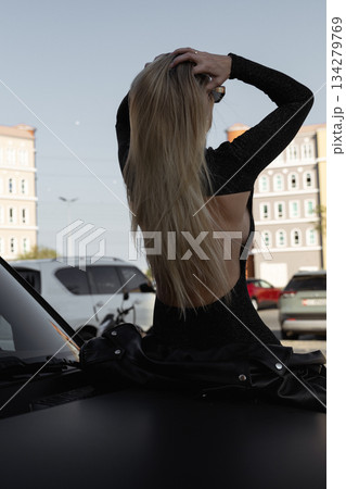 A beautiful girl with blonde hair sits on the hood of a large black car 134279769