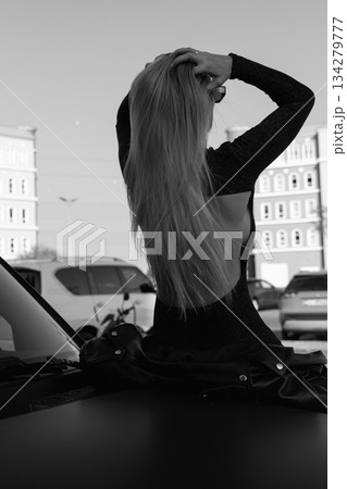 A beautiful girl with blonde hair sits on the hood of a large black car 134279777