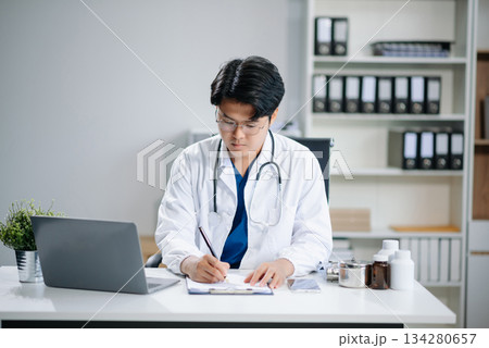 Confident young Asian male doctor in white medical uniform sit at desk working on computer. Smiling use laptop write Confident young Asian male doctor in white medical uniform sit at desk working on computer. Smiling use laptop write 134280657