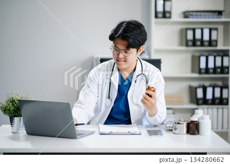 Confident young Asian male doctor in white medical uniform sit at desk working on computer. Smiling use laptop write Confident young Asian male doctor in white medical uniform sit at desk working on computer. Smiling use laptop write 134280662