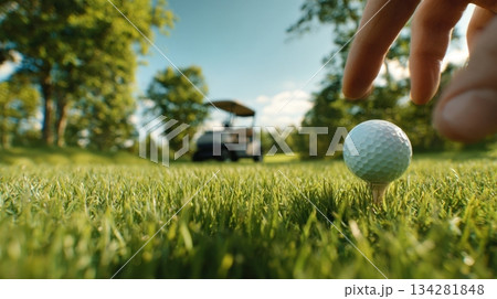 Close up of a golf ball on tee as golfer reaches down on a sunny green golf course Close up of a golf ball on tee as golfer reaches down on a sunny green golf course 134281848
