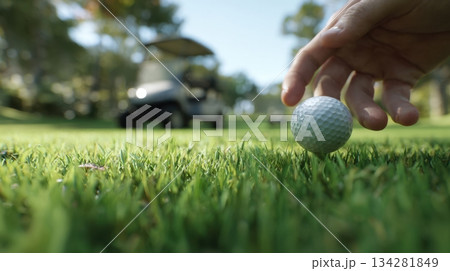 Close up of a golf ball on tee as golfer reaches down on a sunny green golf course 134281849