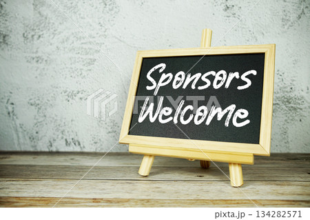 wooden easel displays a blackboard with the bold white text "Sponsors Welcome" set against a rustic wooden surface and textured gray wall background 134282571