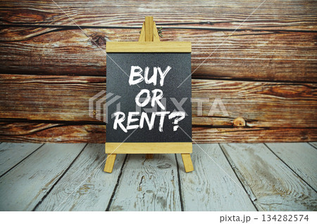 wooden easel holds a blackboard with the white text "Buy or Rent?" set against a rustic wooden wall and light-colored wooden floor 134282574