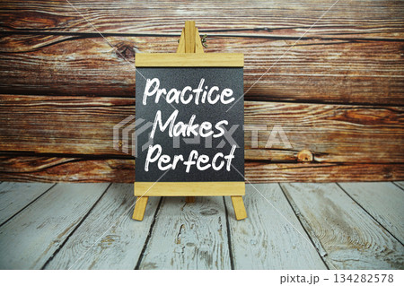 wooden easel holds a blackboard with the white text "Practice Makes Perfect" set against a rustic wooden wall and light-colored wooden floor 134282578