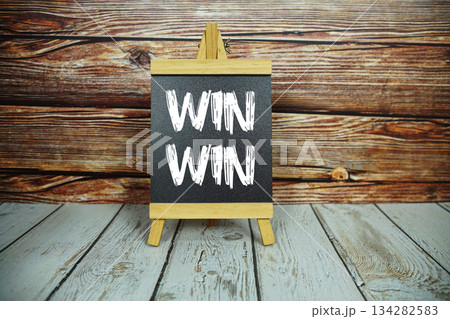 wooden easel holds a blackboard with the white text "Win-Win" set against a rustic wooden wall and light-colored wooden floor 134282583