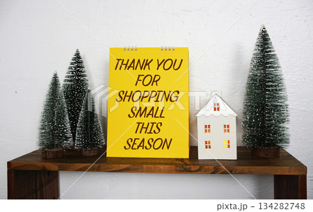 A bright yellow sign with the message "Thank You For Shopping Small This Season" sits on a wooden shelf, surrounded by miniature pine trees and a small decorative white house, creating a festive and 134282748