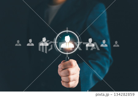 Businessman holding magnifying glass focusing on candidate icon, symbolizing recruitment, human resources, employee selection, target audience, and business hiring strategy. 134282859