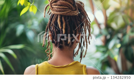 A young African woman with dreadlocks is seen from behind, wearing a yellow tank top. She is surrounded by lush green plants in a natural setting. 134282950
