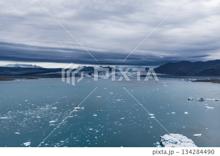 Aerial view of Jokulsarlon Glacier Lagoon in Iceland, featuring blue waters with icebergs, a massive glacier, dark mountains, and a small boat. Aerial view of Jokulsarlon Glacier Lagoon in Iceland, featuring blue waters with icebergs, a massive glacier, dark mountains, and a small boat. 134284490