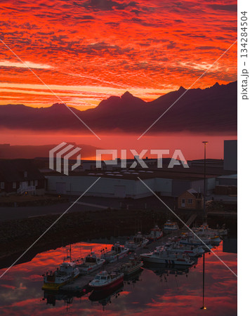 A vibrant sunset reflects on calm harbor waters in Iceland, with docked boats in the foreground and misty jagged mountains in the background. 134284504