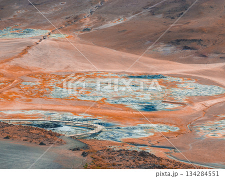 Geothermal area in Iceland with orange and blue hues, steaming vents, mud pots, and a wooden walkway winding through the rugged landscape. 134284551