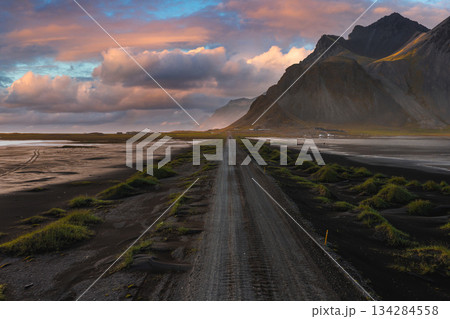A gravel road winds through black sand toward jagged mountains under a vibrant sunset sky, likely on the Stokknes Peninsula in Iceland. 134284558