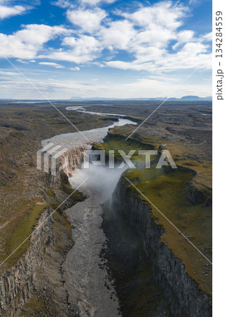 Aerial view of Dettifoss waterfall in Iceland, surrounded by rugged cliffs, a dramatic canyon, rocky terrain, green moss, and a winding river. Aerial view of Dettifoss waterfall in Iceland, surrounded by rugged cliffs, a dramatic canyon, rocky terrain, green moss, and a winding river. 134284559