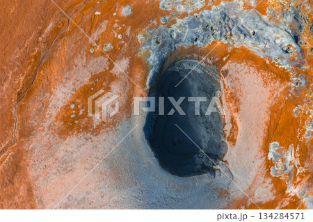Aerial image of a geothermal area in Iceland with orange and gray tones, featuring a dark oval mud pot, mineral deposits, and steaming earth. Aerial image of a geothermal area in Iceland with orange and gray tones, featuring a dark oval mud pot, mineral deposits, and steaming earth. 134284571