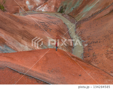 A person in a red jacket stands on a ridge overlooking red and orange toned hills. A winding stream cuts through the rugged, geothermal terrain. 134284585