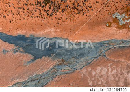 Aerial image of reddish orange geothermal terrain in Iceland, featuring a winding grayish blue stream, textured surface, and scattered vegetation. 134284593