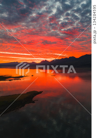 A vivid sunset reflects on calm water in Iceland, with red, orange, and pink hues. Jagged mountains and a grassy peninsula complete the scene. 134284595