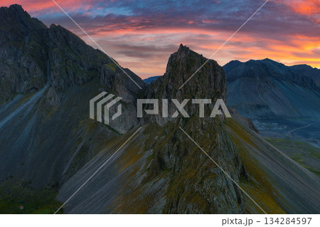 Mountain peaks with a central jagged rock formation at sunset, bathed in orange, pink, and purple hues. Sparse vegetation transitions to rocky terrain. 134284597