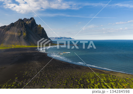 The Stokknes Peninsula in Iceland features sharp mountains, black sand beach, green moss, and calm blue ocean under soft daylight. The Stokknes Peninsula in Iceland features sharp mountains, black sand beach, green moss, and calm blue ocean under soft daylight. 134284598