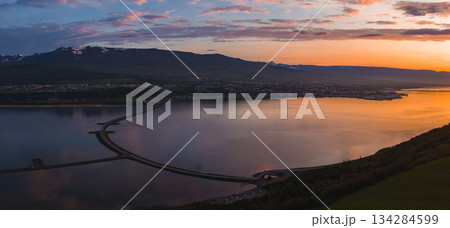 Aerial view of Akureyri, Iceland, at sunset with a calm fjord, a curved bridge crossing the water, and snow capped mountains in the background. Aerial view of Akureyri, Iceland, at sunset with a calm fjord, a curved bridge crossing the water, and snow capped mountains in the background. 134284599