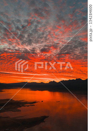 Fiery red and orange sunset reflects on calm water with jagged mountain silhouettes in the background, under a sky filled with textured clouds. 134284600